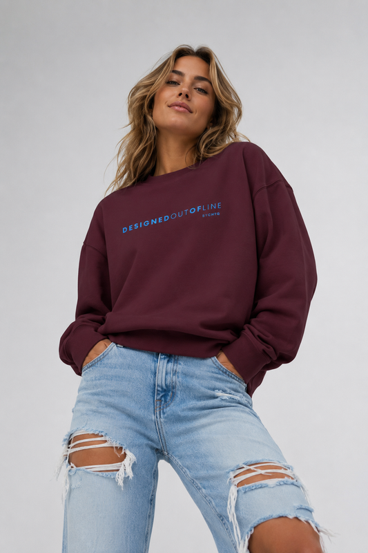 A woman wearing a maroon crewneck sweatshirt with “DESIGNED OUT OF LINE” printed in blue across the chest, paired with light-wash ripped jeans, photographed against a neutral light gray studio background.