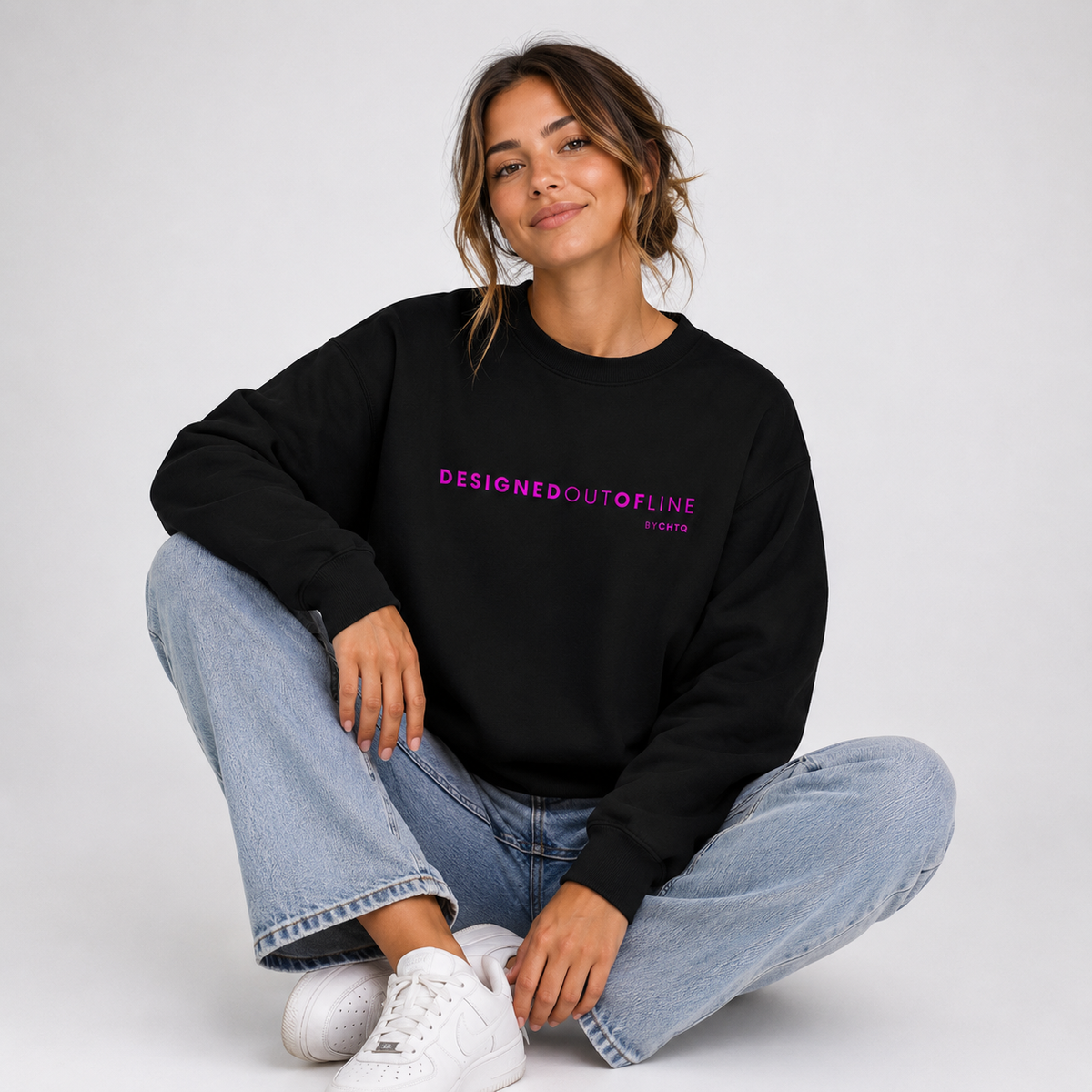 A woman sitting cross-legged against a light gray studio background, wearing a black crewneck sweatshirt with “DESIGNED OUT OF LINE” printed in bright pink across the chest, paired with light-wash blue jeans and white sneakers.