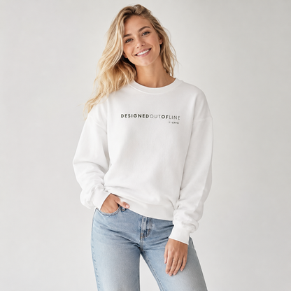 A woman wearing a white crewneck sweatshirt with “DESIGNED OUT OF LINE” printed in dark green across the chest, paired with light-wash jeans, photographed against a neutral light gray studio background.