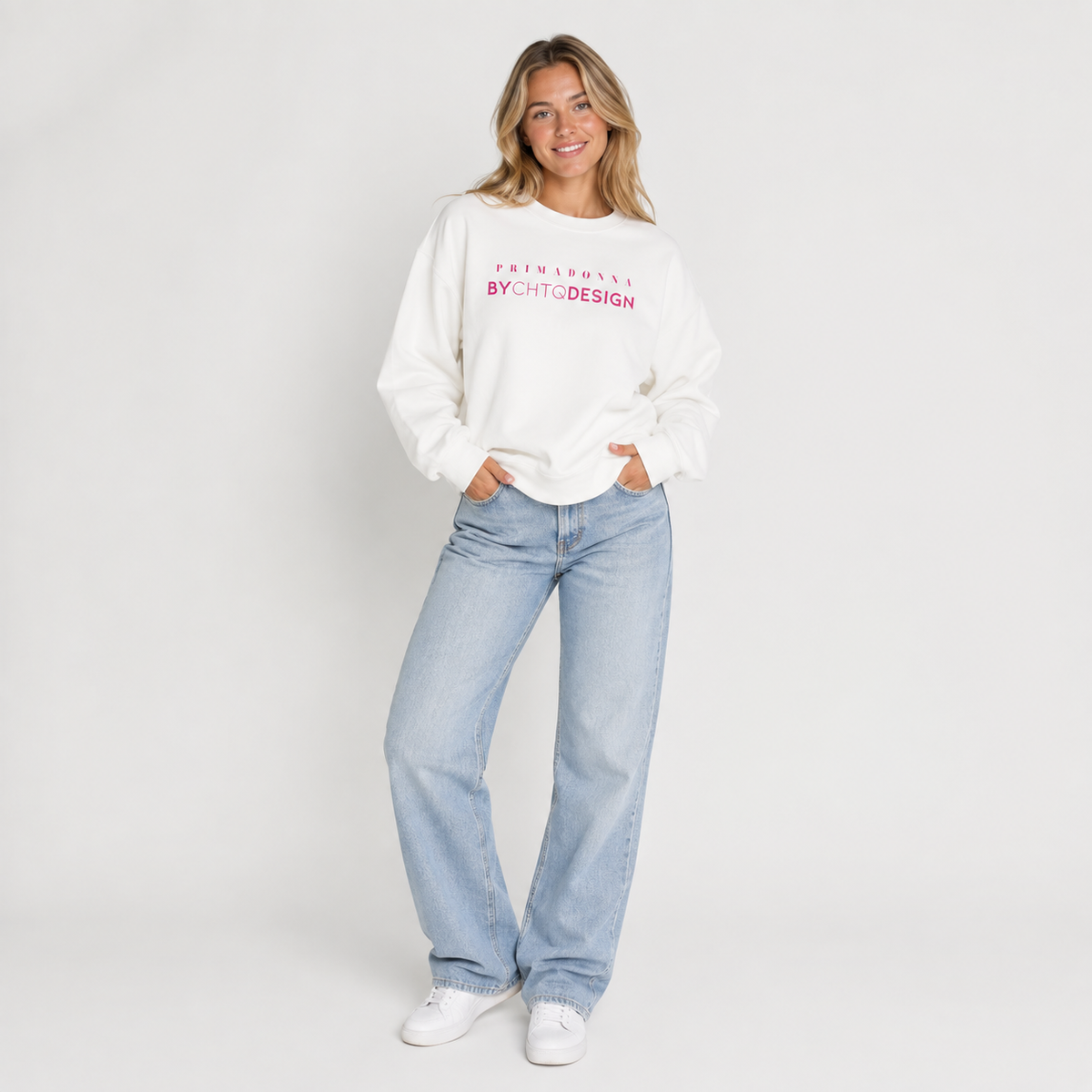 A woman wearing a white crewneck sweatshirt featuring pink “PRIMADONNA BY CHTQ DESIGN” text across the chest, paired with jeans, photographed against a light gray studio background.