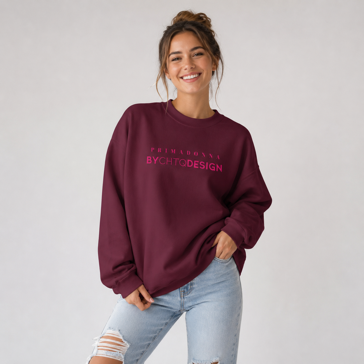 A woman wearing a maroon crewneck sweatshirt featuring pink “PRIMADONNA BY CHTQ DESIGN” text across the chest, paired with jeans, photographed against a light gray studio background.