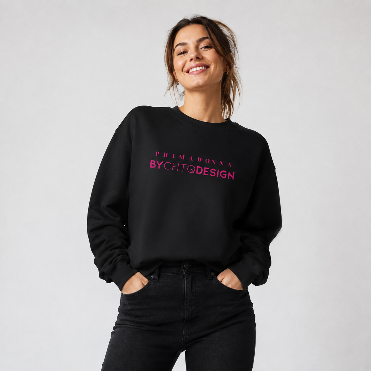 A woman wearing a black crewneck sweatshirt featuring pink “PRIMADONNA BY CHTQ DESIGN” text across the chest, paired with black jeans, photographed against a light gray studio background.