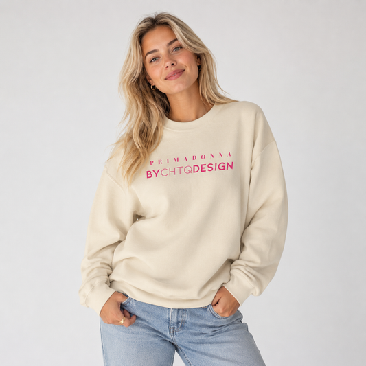 A woman wearing a cream-colored crewneck sweatshirt featuring pink “PRIMADONNA BY CHTQ DESIGN” text across the chest, paired with light blue jeans, photographed against a plain light gray studio background.
