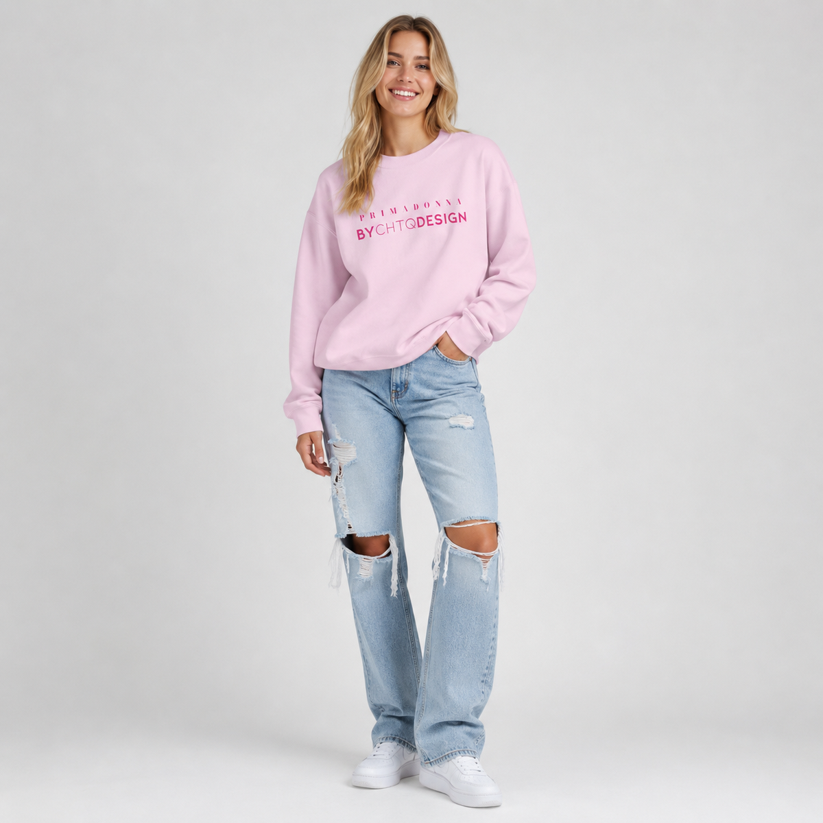 A woman modeling a light pink crewneck sweatshirt featuring pink “PRIMADONNA BY CHTQ DESIGN” text across the chest, paired with distressed light blue jeans and white sneakers, photographed against a light gray studio background.