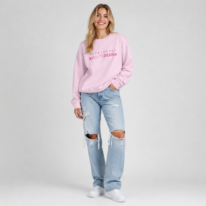 A woman modeling a light pink crewneck sweatshirt featuring pink “PRIMADONNA BY CHTQ DESIGN” text across the chest, paired with distressed light blue jeans and white sneakers, photographed against a light gray studio background.