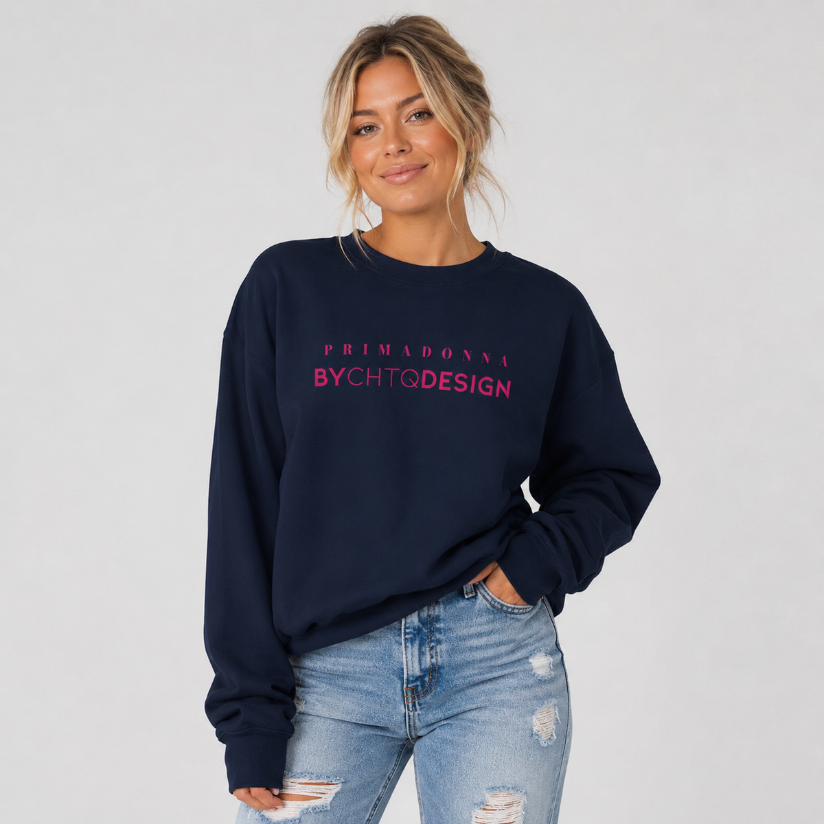 A woman wearing a navy blue crewneck sweatshirt featuring pink “PRIMADONNA BY CHTQ DESIGN” text across the chest, paired with light blue jeans, photographed against a plain light gray studio background.