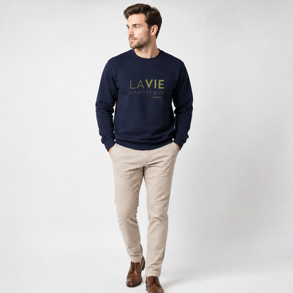 A man wearing a navy blue LA VIE CHAOTIQUE graphic crewneck sweatshirt with olive green lettering, paired with beige trousers and dark brown shoes against a light gray studio background.