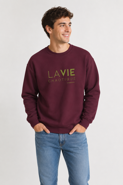 A man wearing a maroon LA VIE CHAOTIQUE graphic crewneck sweatshirt with olive green lettering, paired with beige trousers and dark brown shoes against a light gray studio background.