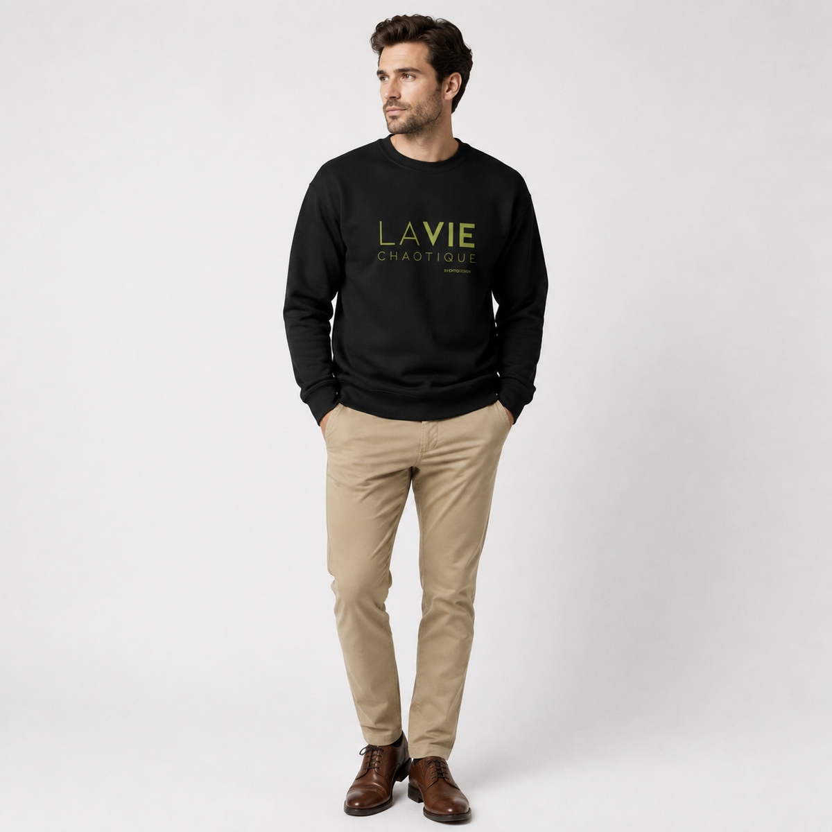 A man wearing a black LA VIE CHAOTIQUE graphic crewneck sweatshirt with olive green lettering, paired with beige trousers and dark brown shoes against a light gray studio background.