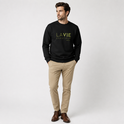 A man wearing a black LA VIE CHAOTIQUE graphic crewneck sweatshirt with olive green lettering, paired with beige trousers and dark brown shoes against a light gray studio background.