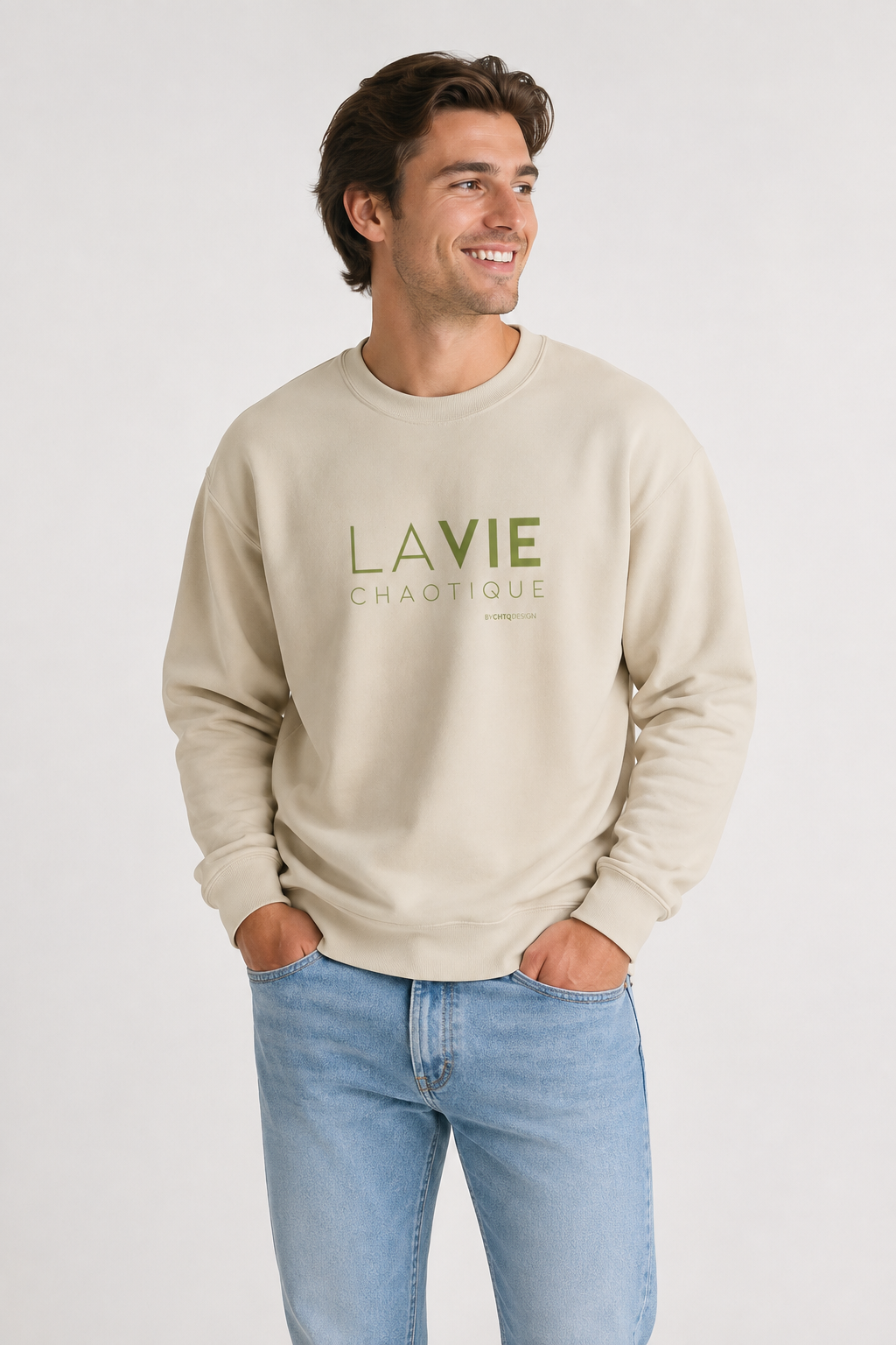 A man wearing a beige LA VIE CHAOTIQUE graphic crewneck sweatshirt with olive green lettering, paired with beige trousers and dark brown shoes against a light gray studio background.