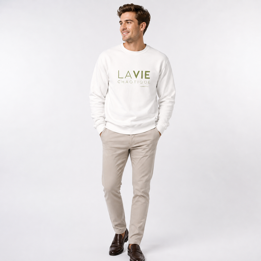 A man wearing a white LA VIE CHAOTIQUE graphic crewneck sweatshirt with olive green lettering, paired with beige trousers and dark brown shoes against a light gray studio background.