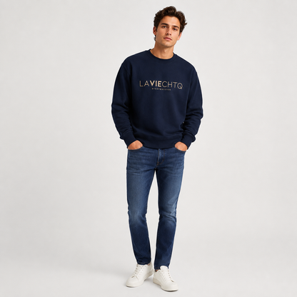 A man wearing a navy crewneck sweatshirt with “LA VIE CHTQ” and “BY CHTQ DESIGN” printed in gold across the chest, paired with blue jeans and white sneakers, photographed against a clean white studio background.