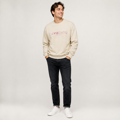 A man wearing a beige crewneck sweatshirt with “LA VIE CHTQ” and “BY CHTQ DESIGN” printed in gold across the chest, paired with blue jeans and white sneakers, photographed against a clean white studio background.
