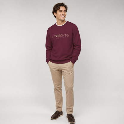 A man wearing a maroon crewneck sweatshirt with “LA VIE CHTQ” and “BY CHTQ DESIGN” printed in gold across the chest, paired with blue jeans and white sneakers, photographed against a clean white studio background.