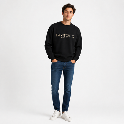 A man wearing a black crewneck sweatshirt with “LA VIE CHTQ” and “BY CHTQ DESIGN” printed in gold across the chest, paired with blue jeans and white sneakers, photographed against a clean white studio background.