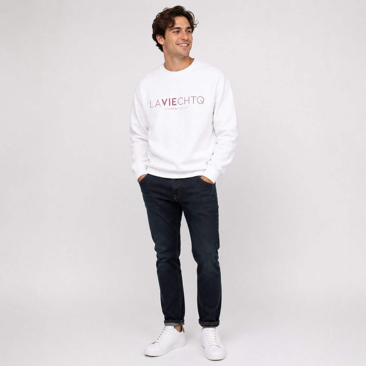 A man wearing a white crewneck sweatshirt with “LA VIE CHTQ” and “BY CHTQ DESIGN” printed in gold across the chest, paired with blue jeans and white sneakers, photographed against a clean white studio background.