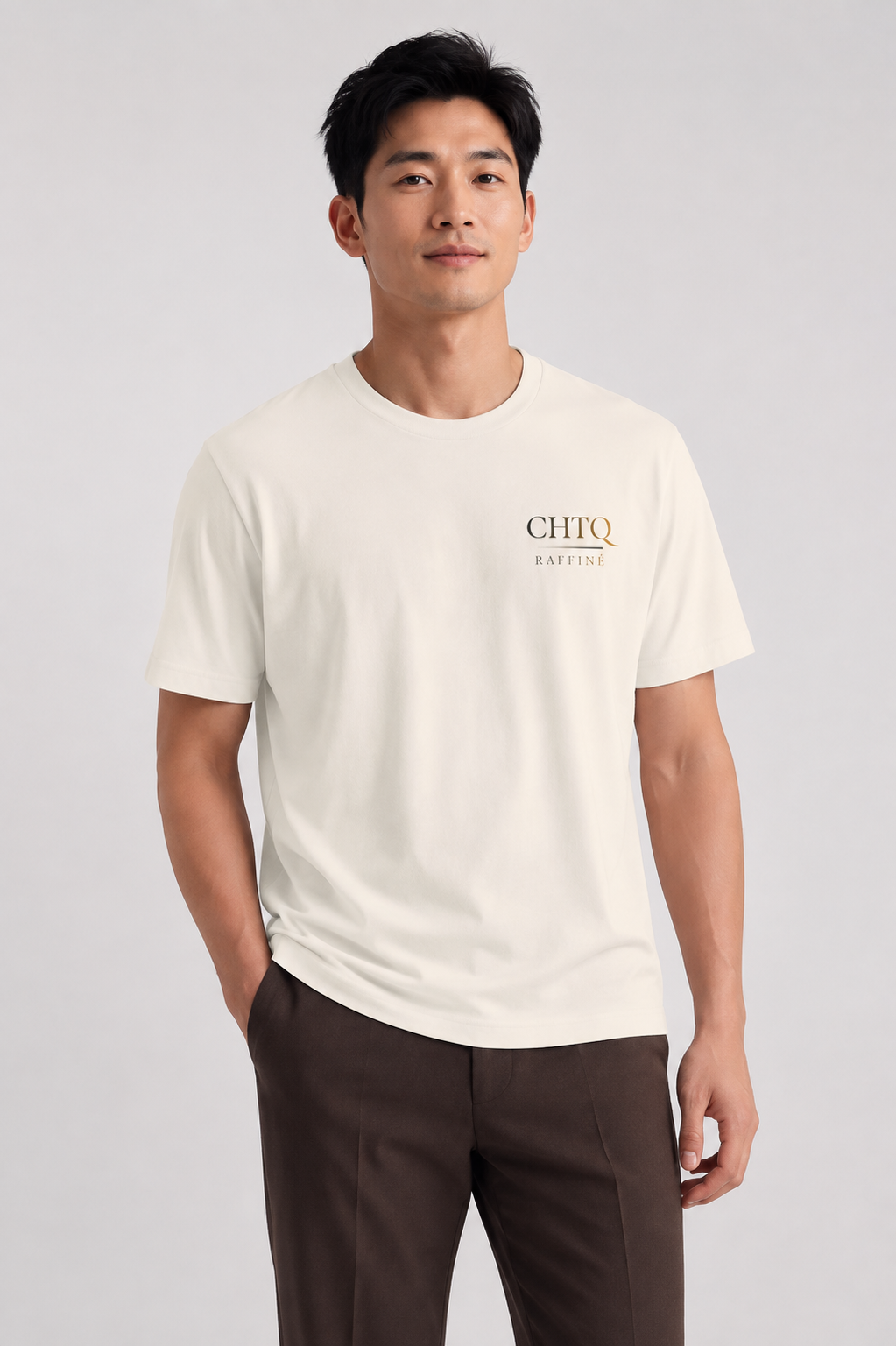 A man wearing a cream-colored short-sleeve t-shirt with a small gold and black 'CHTQ Raffine' logo on the left chest, paired with dark brown trousers, against a light gray studio background.