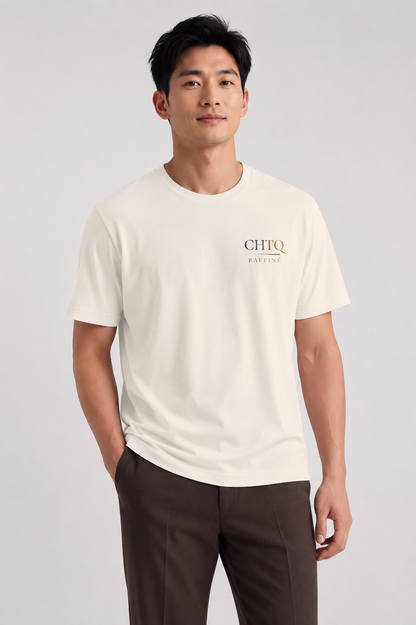 A man wearing a cream-colored short-sleeve t-shirt with a small gold and black 'CHTQ Raffine' logo on the left chest, paired with dark brown trousers, against a light gray studio background.