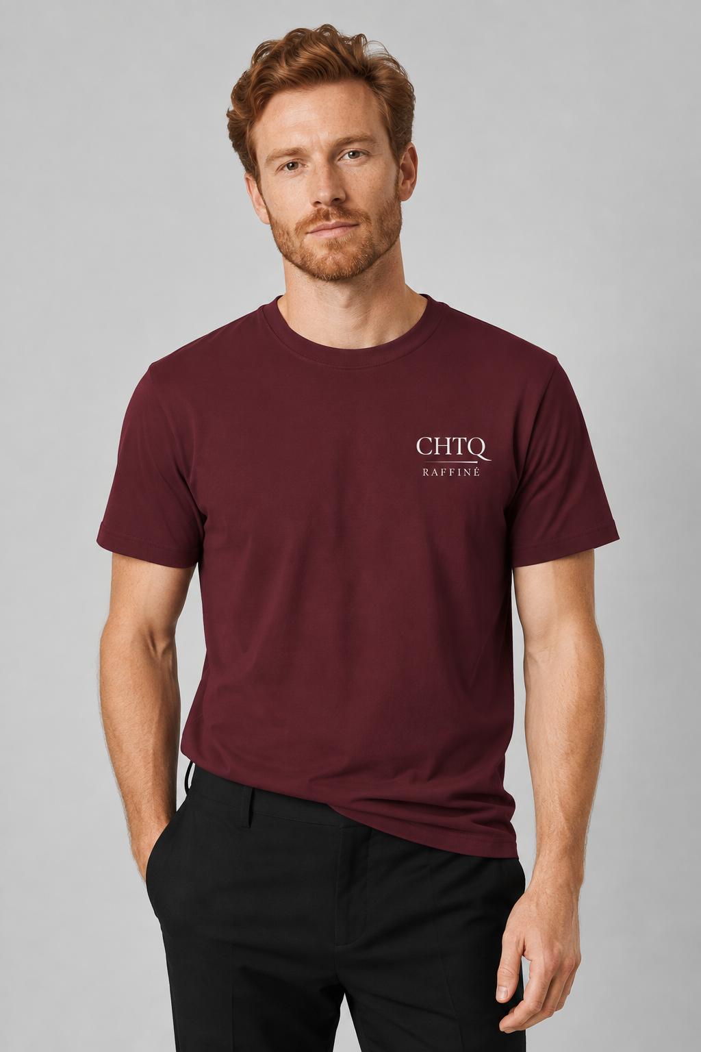 A man wearing a maroon short-sleeve t-shirt with a white 'CHTQ RAFFINE' logo on the left chest, paired with black trousers, against a solid gray studio background.