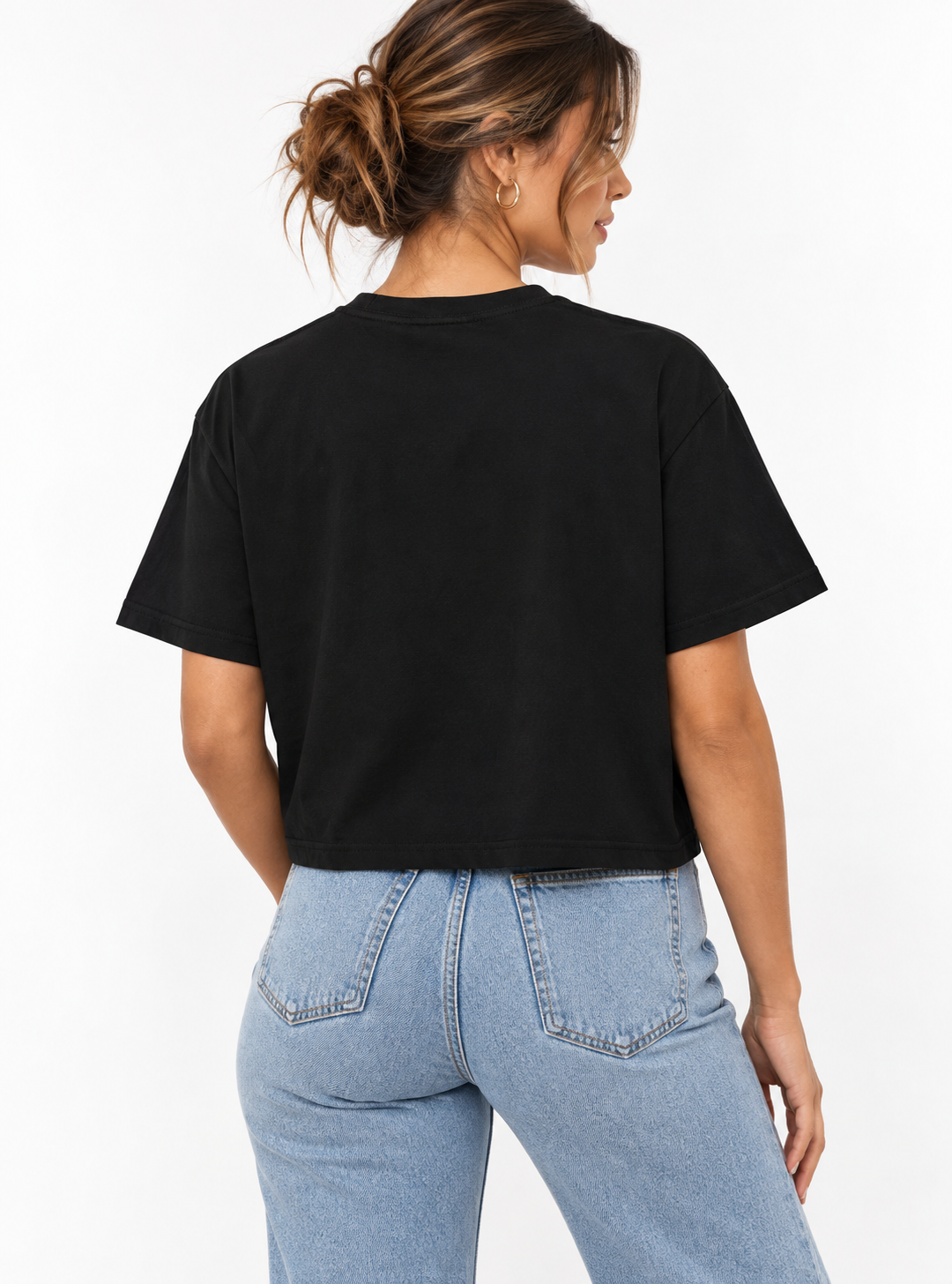 A woman shown from the back wearing a black cropped boxy short-sleeve T-shirt with a gold “DESIGNED OUT OF LINE” typography graphic on the front, paired with light-wash blue jeans against a clean white studio background.