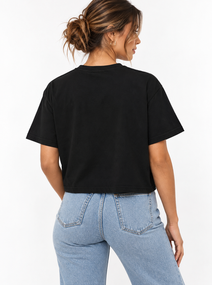 A woman shown from the back wearing a black cropped boxy short-sleeve T-shirt with a gold “DESIGNED OUT OF LINE” typography graphic on the front, paired with light-wash blue jeans against a clean white studio background.