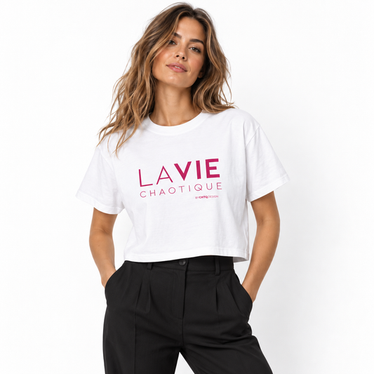 A woman wearing a white LA VIE CHAOTIQUE cropped boxy T-shirt with pink typography on the front, paired with high-waisted black trousers against a clean white studio background.