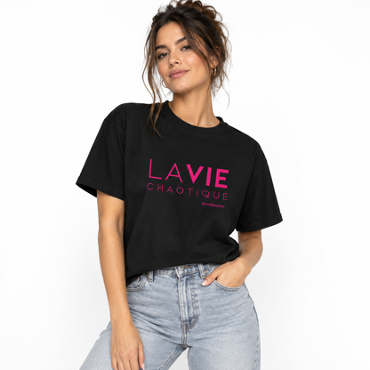 A woman wearing a black LA VIE CHAOTIQUE graphic boxy T-shirt with pink and white typography on the front, paired with light-wash blue jeans against a clean white studio background.