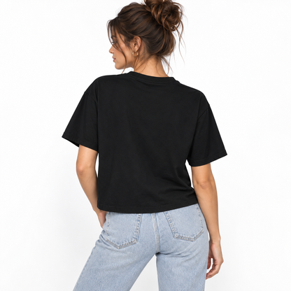 A woman shown from the back wearing a black LA VIE CHAOTIQUE cropped boxy T-shirt with pink typography on the front, paired with light-wash blue jeans against a clean white studio background.