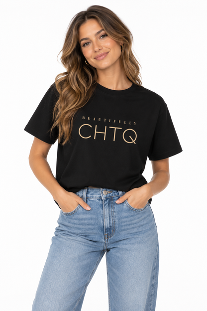 A woman modeling a black short-sleeve boxy graphic T-shirt with the text “BEAUTIFULLY CHTQ” printed in gold on the chest, paired with light blue high-waisted jeans against a plain white studio background.