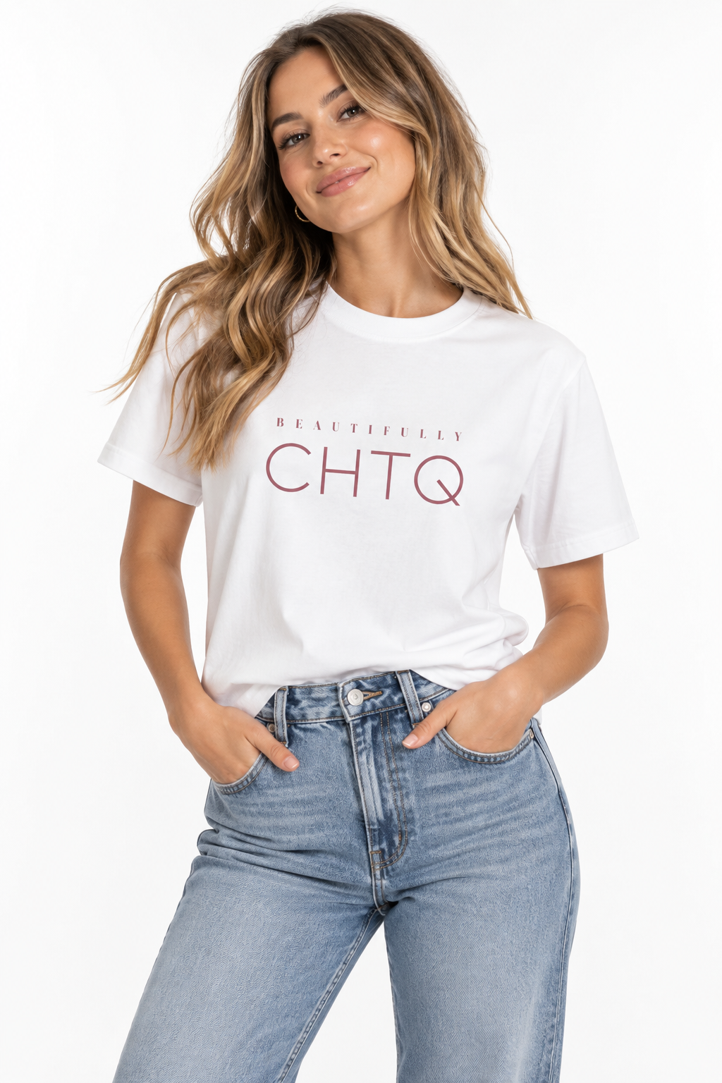 A woman wearing a white boxy graphic T-shirt with “BEAUTIFULLY CHTQ” printed in dusty pink on the front, paired with light-wash high-waisted jeans against a clean white studio background.