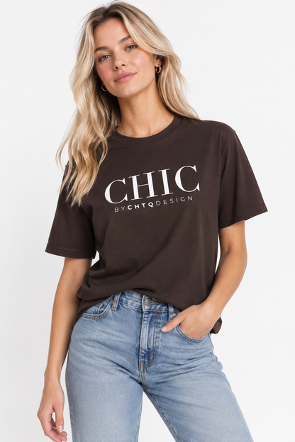 A woman wearing a brown CHIC by CHTQ Design graphic T-shirt with white typography, paired with light blue jeans against a clean white studio background.