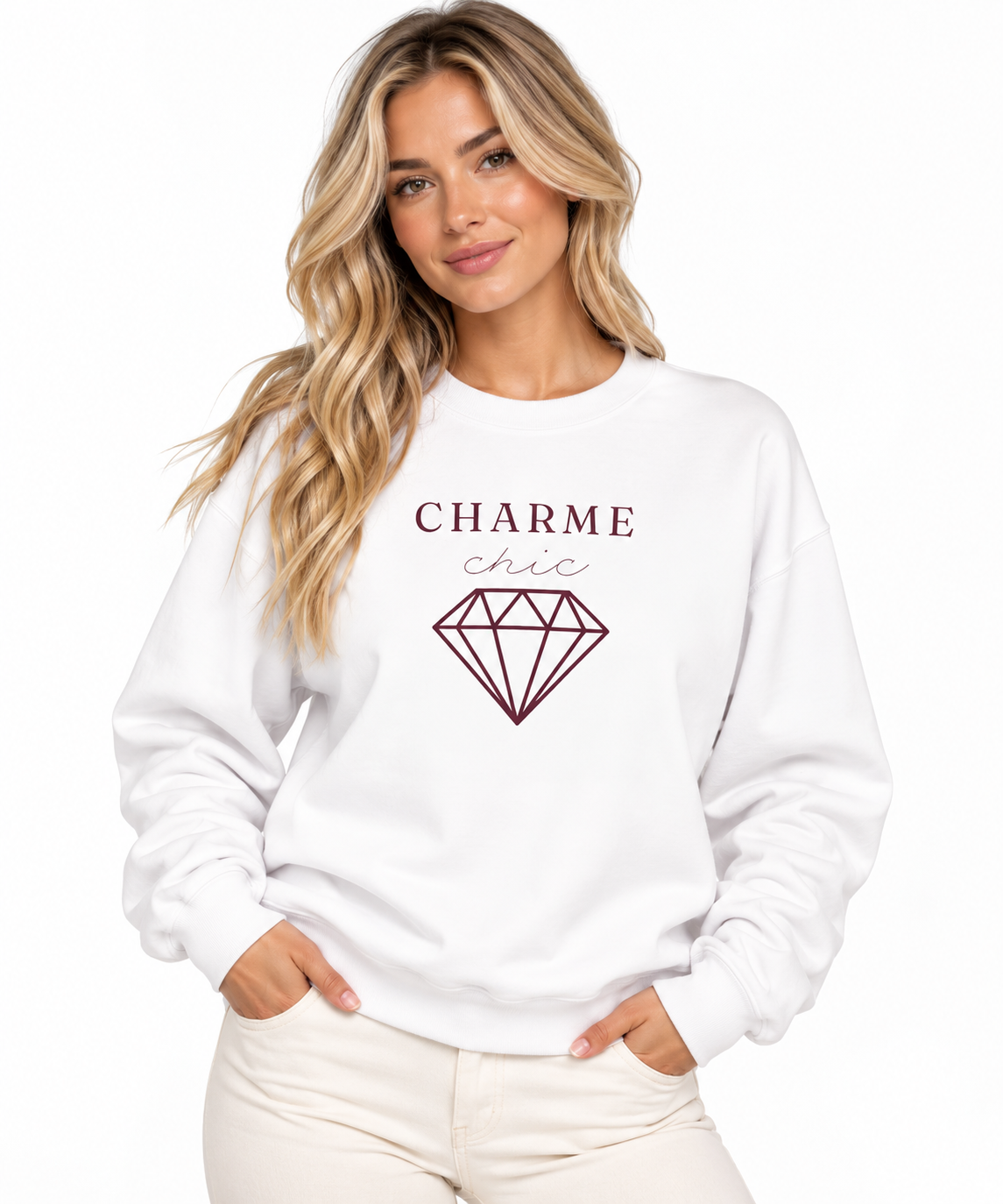 A woman modeling a white crewneck sweatshirt featuring a burgundy “CHARME chic” graphic with a diamond outline, paired with cream pants against a clean white studio background.
