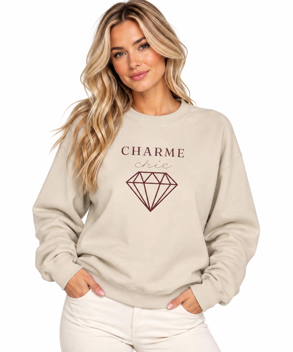 A woman modeling a beige crewneck sweatshirt featuring a burgundy “CHARME chic” graphic with a diamond outline, paired with cream pants against a clean white studio background.