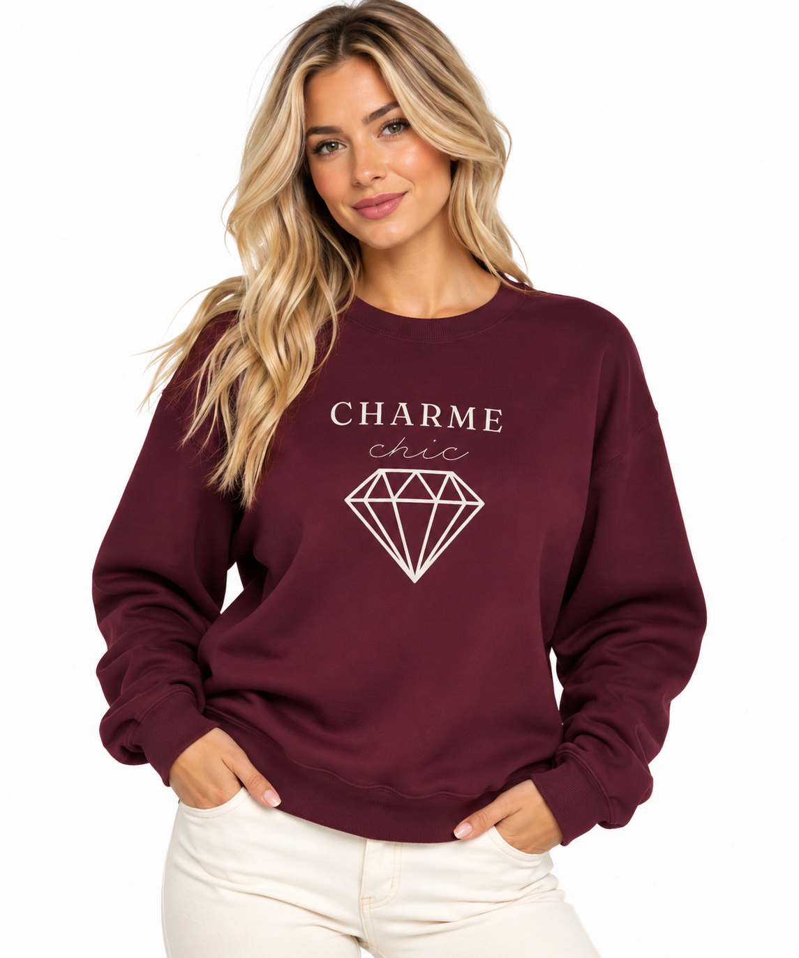 A woman modeling a maroon crewneck sweatshirt featuring a light “CHARME chic” graphic with a diamond outline, paired with cream pants against a clean white studio background.