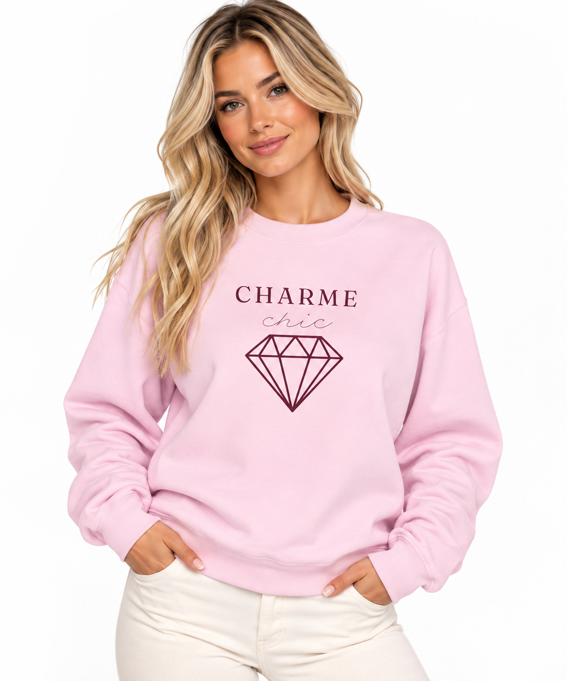 A woman modeling a light pink crewneck sweatshirt featuring a burgundy “CHARME chic” graphic with a diamond outline, paired with cream pants against a clean white studio background.
