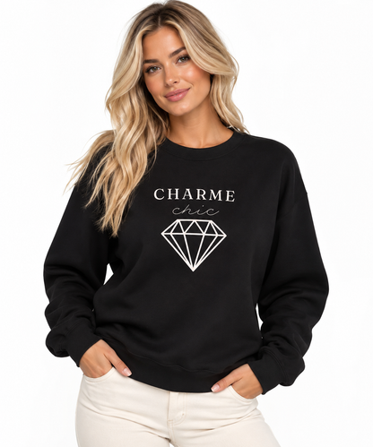 A woman modeling a black crewneck sweatshirt featuring a light “CHARME chic” graphic with a diamond outline, paired with cream pants against a clean white studio background.