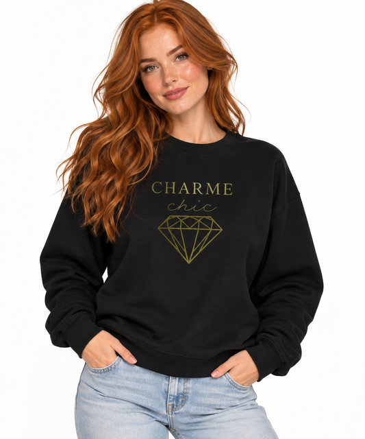 A woman with long red hair wearing a black crewneck sweatshirt with a gold graphic that reads 'CHARME chic' and a diamond illustration, paired with light blue jeans, against a plain white background.