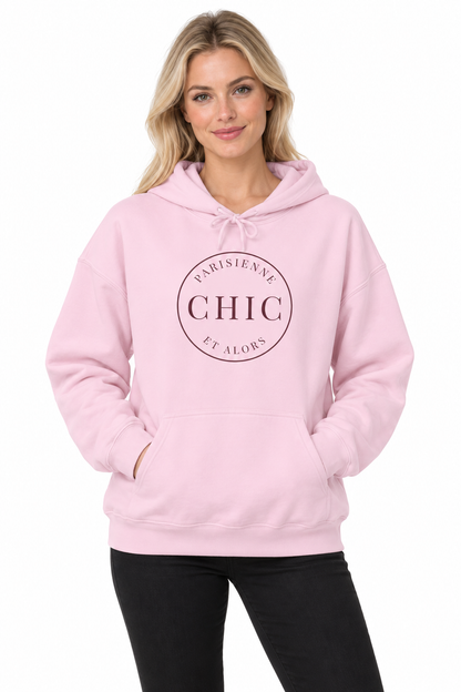 A woman modeling a light pink pullover hoodie with a circular dark pink graphic on the chest reading 'PARISIENNE CHIC ET ALORS'.