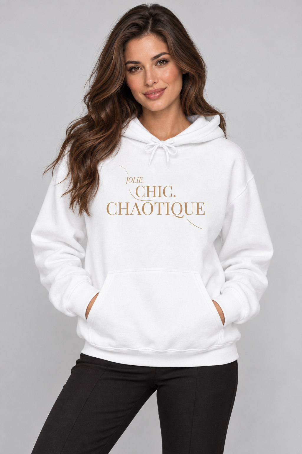 A woman wearing a white pullover hoodie with 'Jolie. CHIC. CHAOTIQUE' printed in gold lettering on the chest, paired with black pants, against a solid gray studio background.