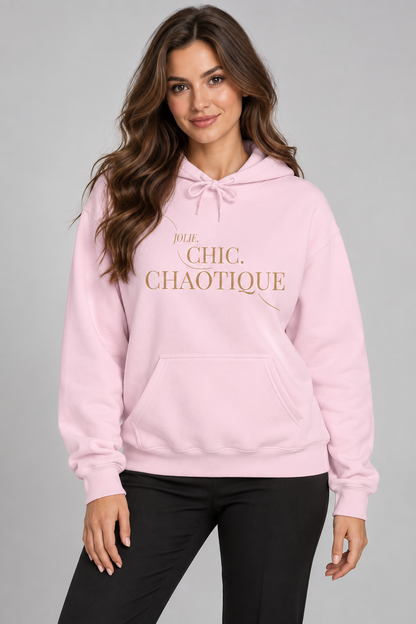 A woman wearing a light pink pullover hoodie with 'Jolie CHIC. CHAOTIQUE' printed in gold lettering on the chest, paired with black pants, against a solid gray studio background.