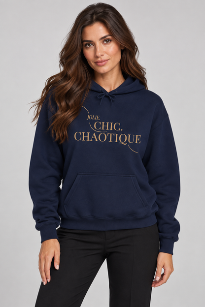 A woman wearing a navy blue hoodie with gold text reading 'Jolie Chic Chaotique' on the chest, paired with black pants, against a light gray studio background.