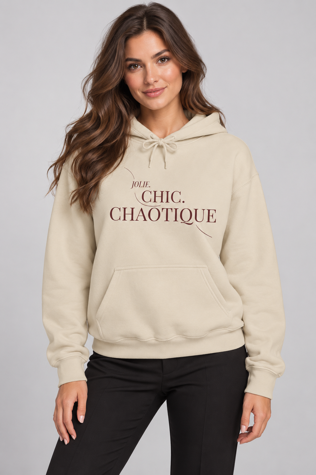 A woman wearing a cream-colored hoodie with the text 'Jolie Chic. Chaotique' printed on the chest, paired with black pants, against a solid gray studio background.