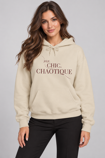 A woman wearing a cream-colored hoodie with the text 'Jolie Chic. Chaotique' printed on the chest, paired with black pants, against a solid gray studio background.