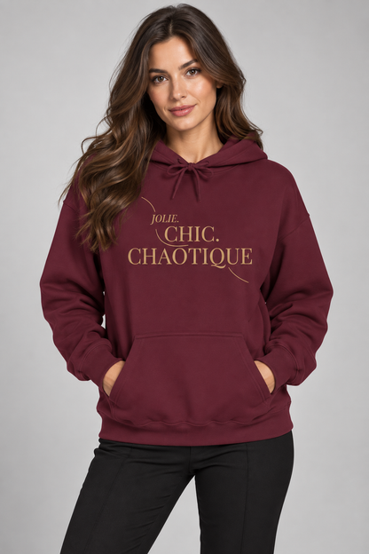 A woman modeling a burgundy hoodie with gold text reading 'Jolie Chic Chaotique' on the chest, paired with black pants, against a neutral gray studio background.