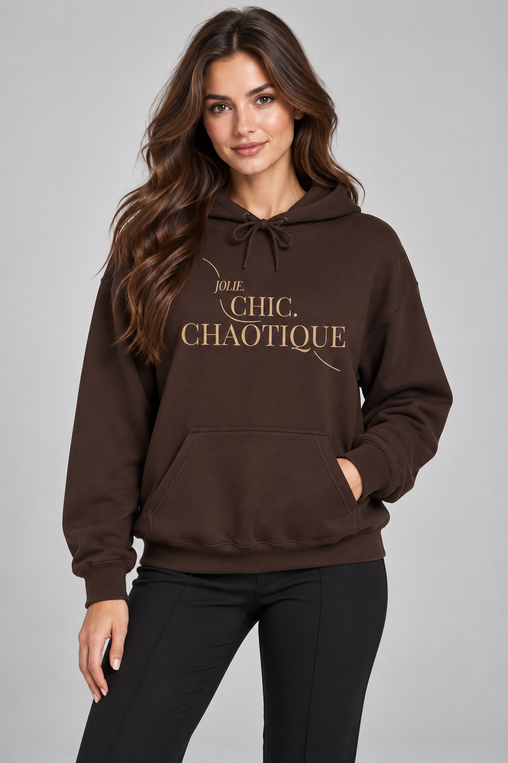 A woman wearing a dark brown pullover hoodie with 'Jolie Chic. Chaotique' printed in gold lettering on the chest, paired with black pants, against a solid gray studio background.