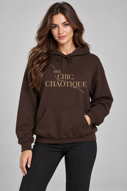 A woman wearing a dark brown pullover hoodie with 'Jolie Chic. Chaotique' printed in gold lettering on the chest, paired with black pants, against a solid gray studio background.