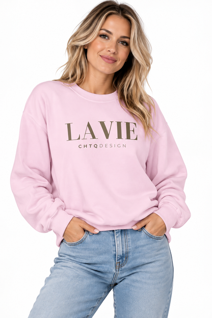 A woman wearing a light pink crewneck sweatshirt with 'LAVIE' and 'CHTQDESIGN' printed on the front, paired with light blue jeans, against a plain white background.