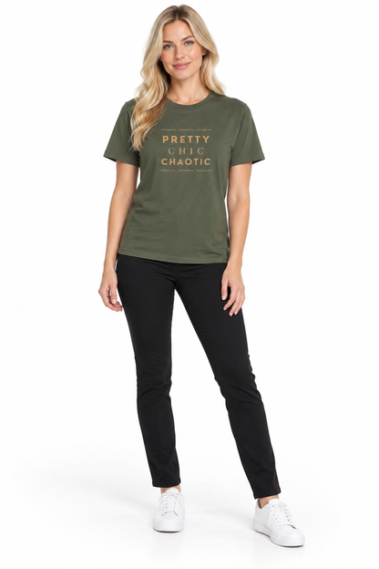 A woman modeling an olive green graphic t-shirt with the words 'PRETTY CHIC Chaotic' printed in gold on the chest, paired with black pants and white sneakers, against a plain white background.