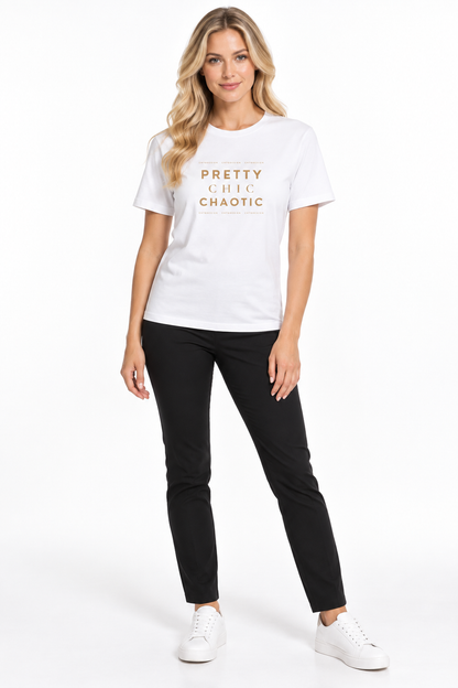A woman modeling a white graphic t-shirt with gold text that reads 'PRETTY CHIC CAOTIC' paired with black pants and white sneakers, set against a plain white studio background.